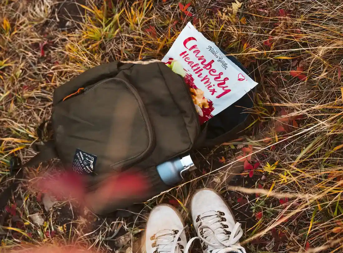 Nature’s Garden Cranberry Health Mix in a hiker’s backpack on dry grass, paired with hiking boots and water bottle for an outdoor snack.