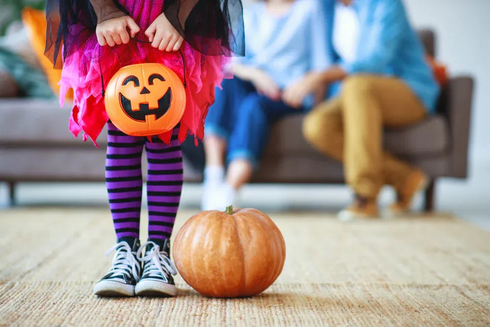 Child in Halloween costume holding a pumpkin trick-or-treat bucket with parents sitting in the background and a pumpkin on the floor.