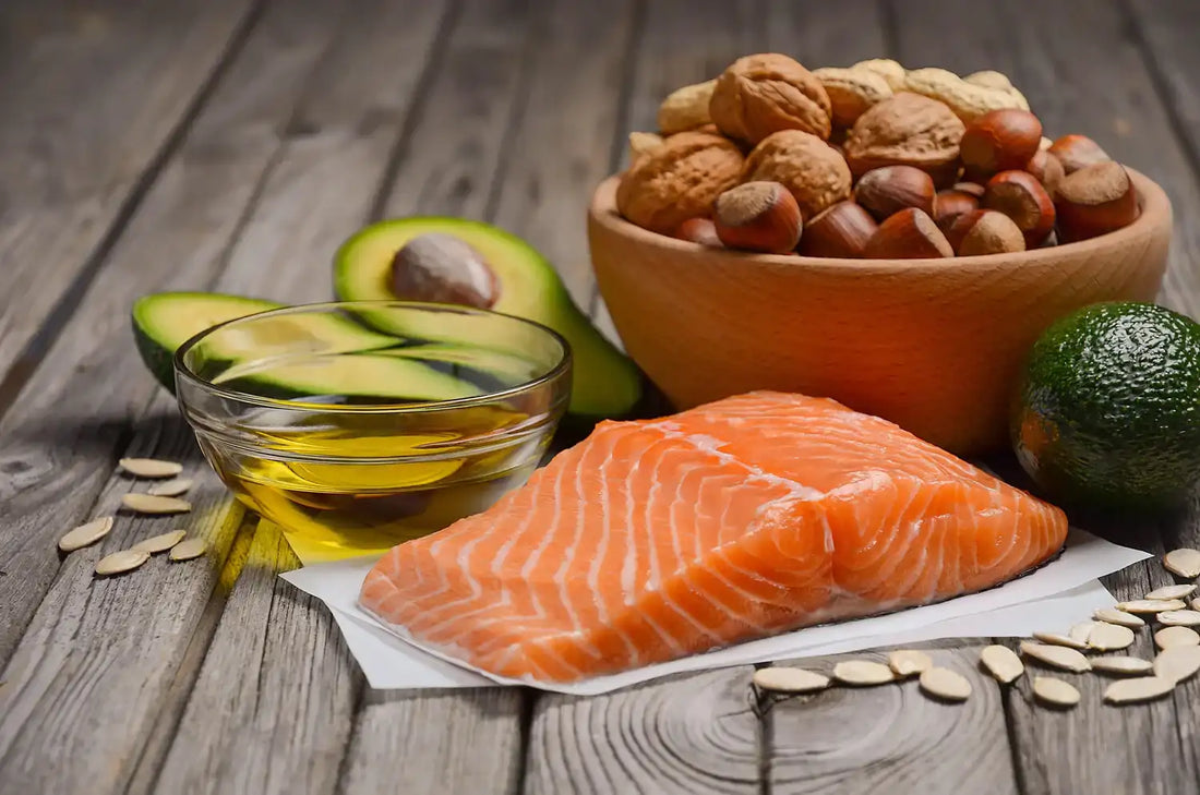 Salmon, avocado, mixed nuts, and olive oil on wood surface—foods rich in healthy fats that support skin and overall wellness.