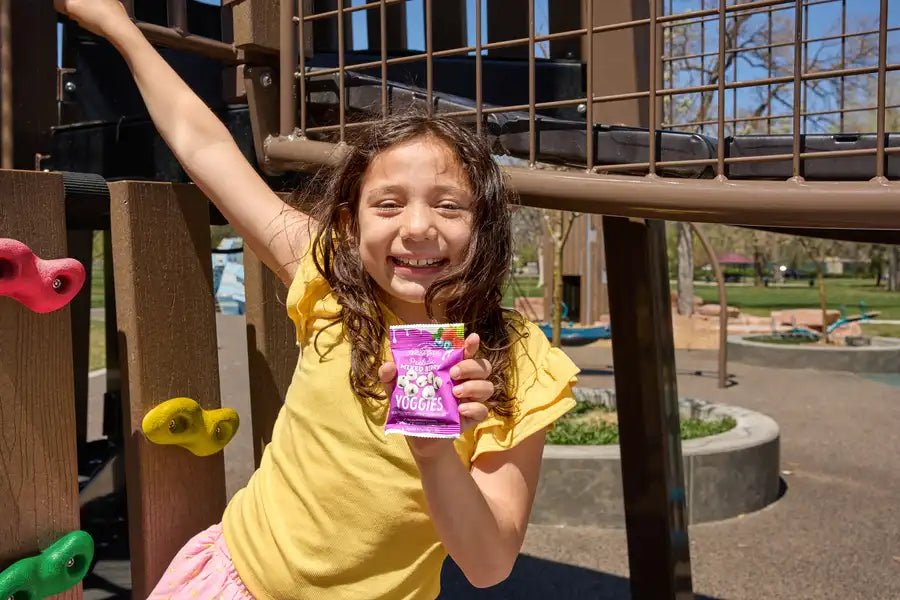 Smiling child at a sunny playground in the USA holding a pack of Nature’s Garden Strawberry Yoggies yogurt-covered fruit bites, a better-for-you probiotic snack for kids.
