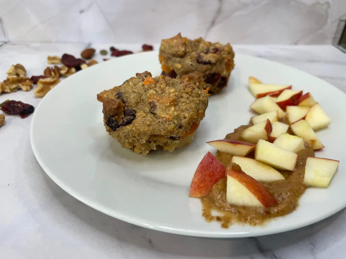 Two homemade morning glory muffins served with chopped apples and almond butter on a white plate, surrounded by dried cranberries and nuts.