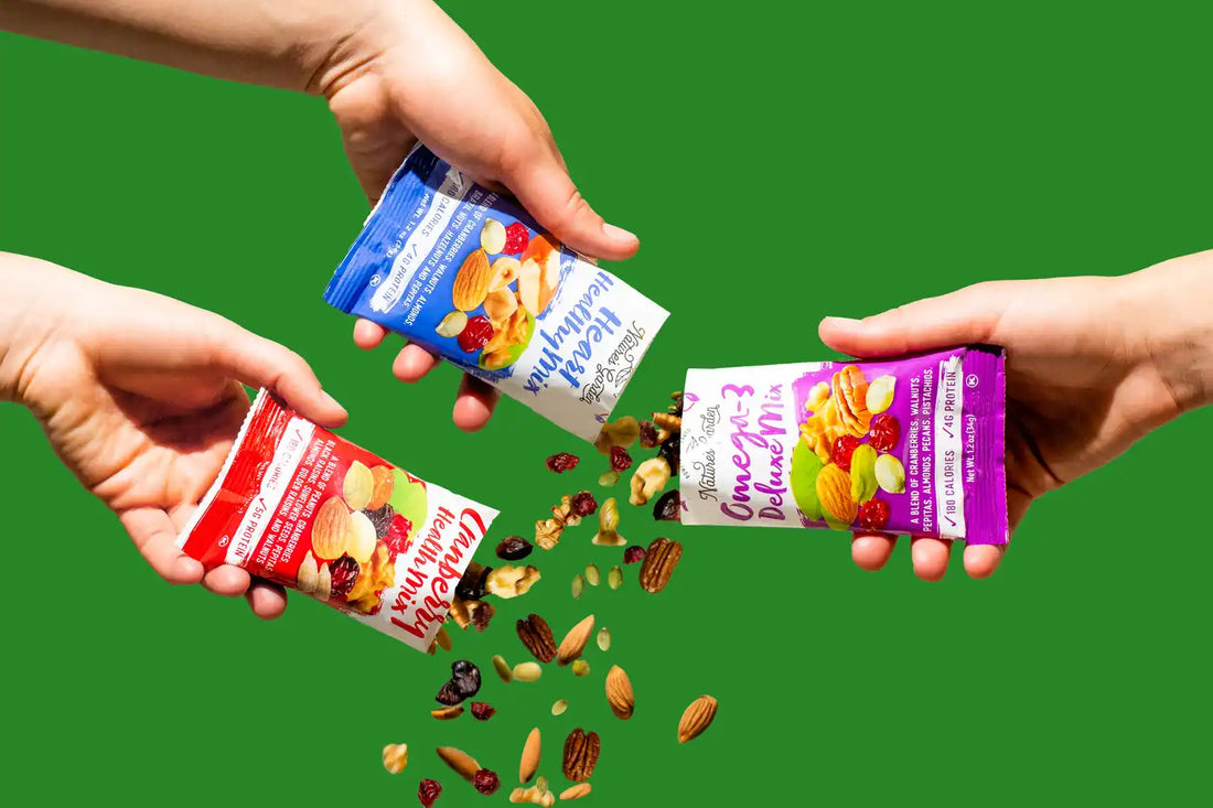 Hands pouring Nature's Garden trail mix pouches—Cranberry Health Mix, Heart Healthy Mix, and Omega-3 Deluxe Mix—against green background
