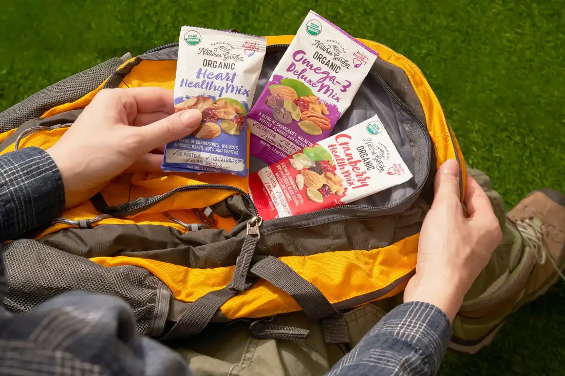 Hands holding Nature’s Garden Organic snack packs inside a hiking backpack, including Heart Healthy, Omega-3, and Cranberry Mix.