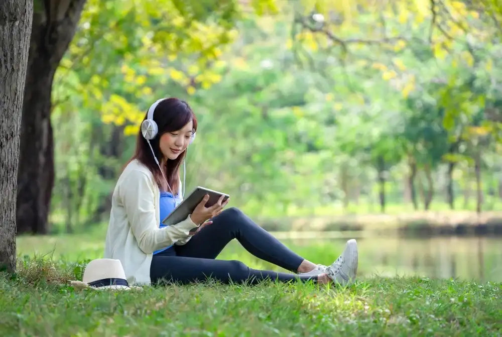 Woman relaxing with headphones and tablet by a lake, enjoying music and summer vibes—perfect pairing for Nature’s Garden snacks