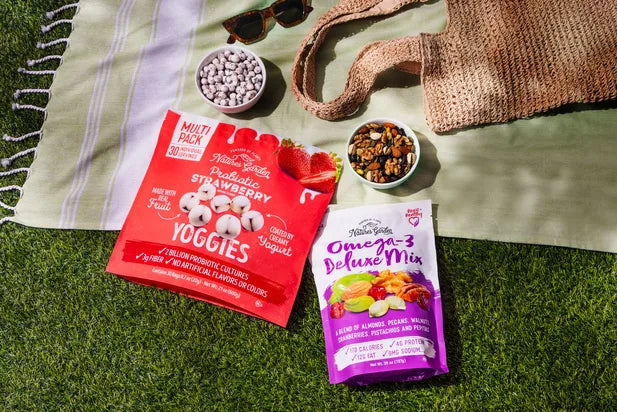 Nature’s Garden Yoggies and Omega-3 Deluxe Mix picnic setup with trail mix, yogurt bites, and snacks on a blanket in sunny grass.