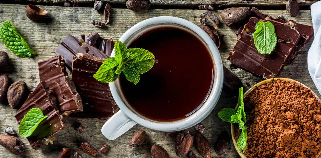 Cup of rich dark chocolate with mint, surrounded by cocoa powder, cacao beans, and sugar-free chocolate pieces on wood.