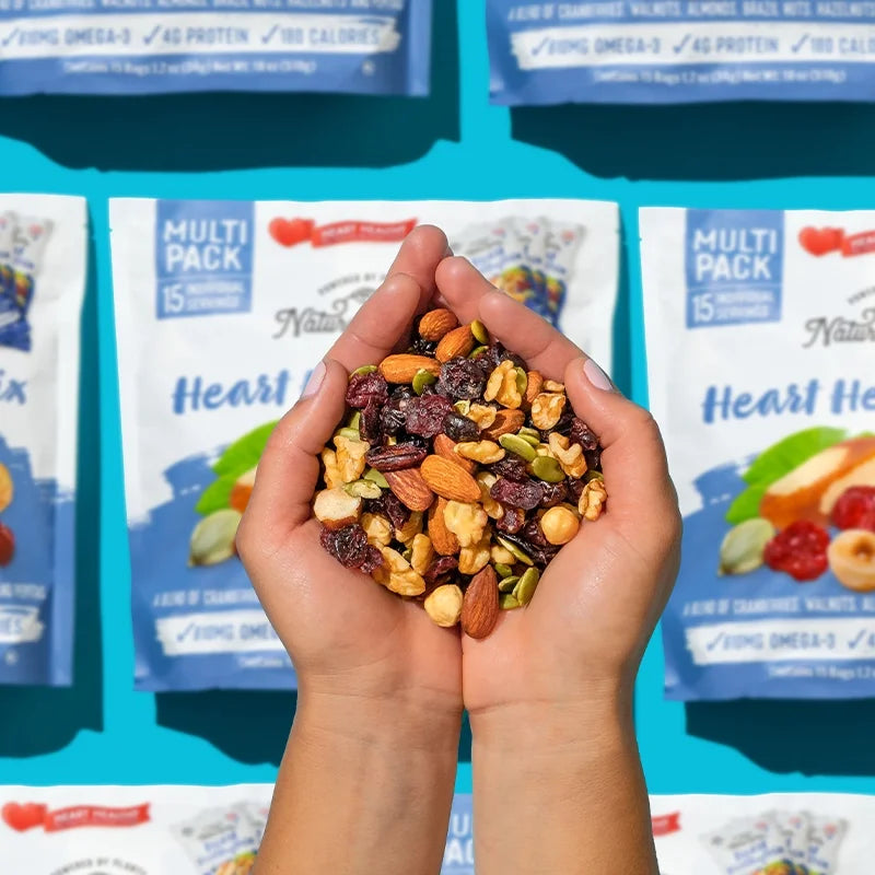 Hands holding Nature’s Garden Heart Healthy Mix with almonds, walnuts, cranberries, and pepitas over branded snack packs on a vibrant blue background.