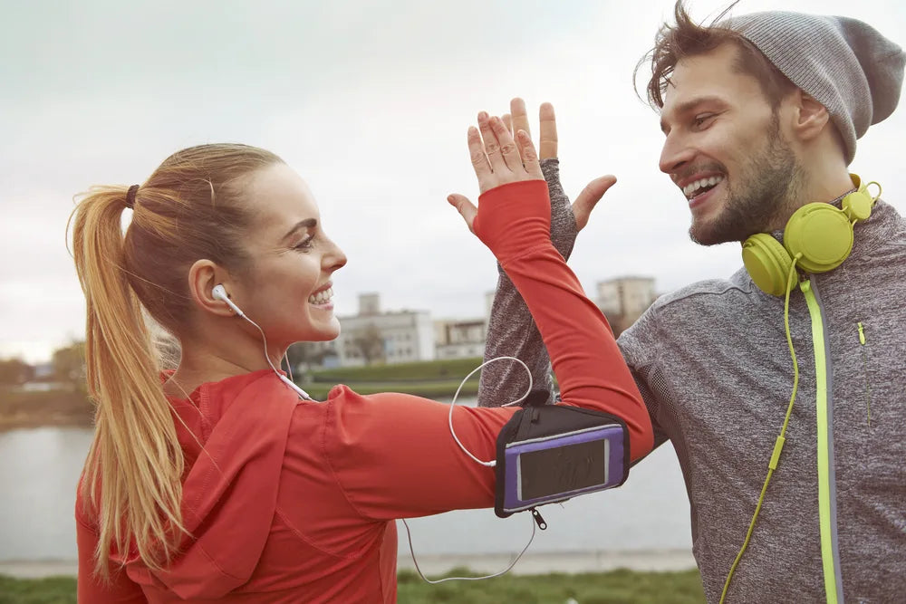 Two active adults in athletic wear smiling and high-fiving after a workout, wearing earbuds and fitness gear outdoors near water.