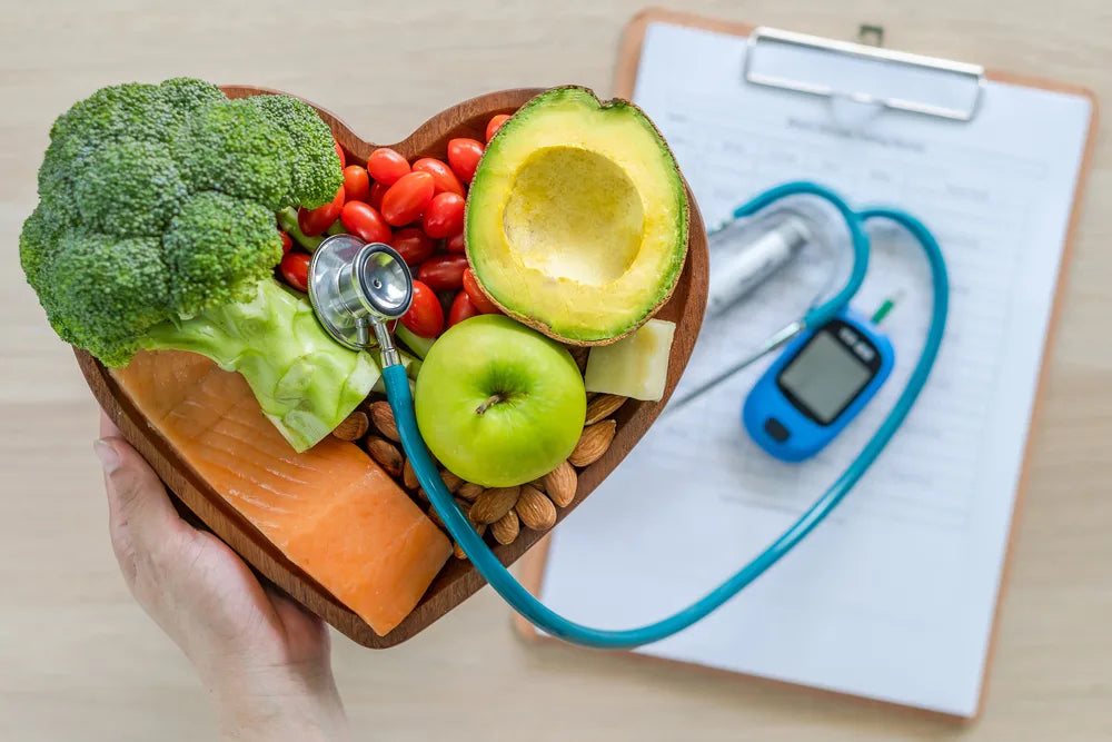 Heart-shaped bowl with salmon, avocado, broccoli, almonds, apple, and tomatoes next to a stethoscope and glucose meter on a clipboard.