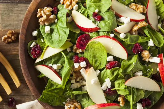 Fresh spinach salad with sliced apples, dried cranberries, walnuts, and feta cheese in a wooden bowl on rustic table.