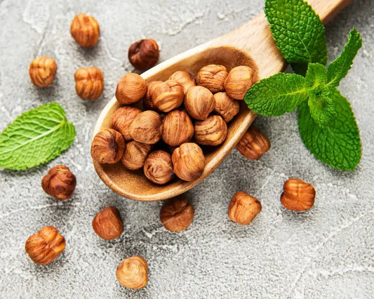 Wooden spoon filled with whole hazelnuts, surrounded by fresh mint leaves on a textured surface for a health-focused blog.