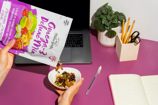 Pouring Omega-3 Deluxe Mix trail mix into a bowl at a pink desk with a laptop, notebook, and office supplies.