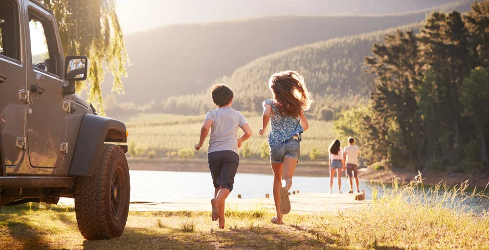 Family enjoying a summer lake getaway with kids running toward the dock near an SUV during golden hour.