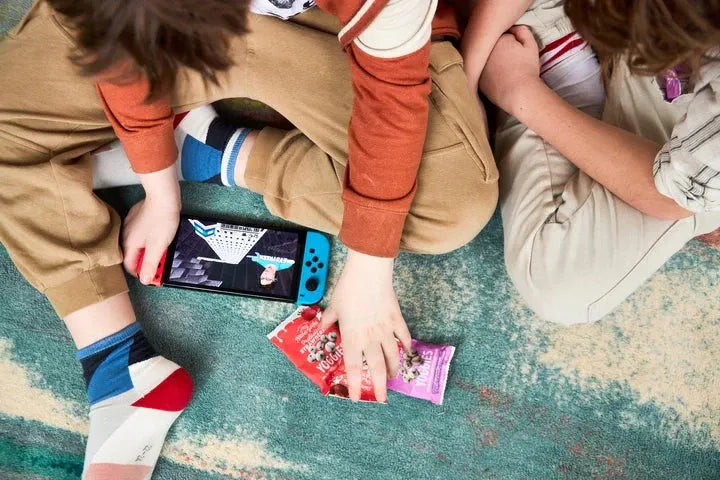 Two kids sitting on a colorful carpet playing a handheld game console while snacking on Nature’s Garden Yoggies yogurt-covered fruit snacks.