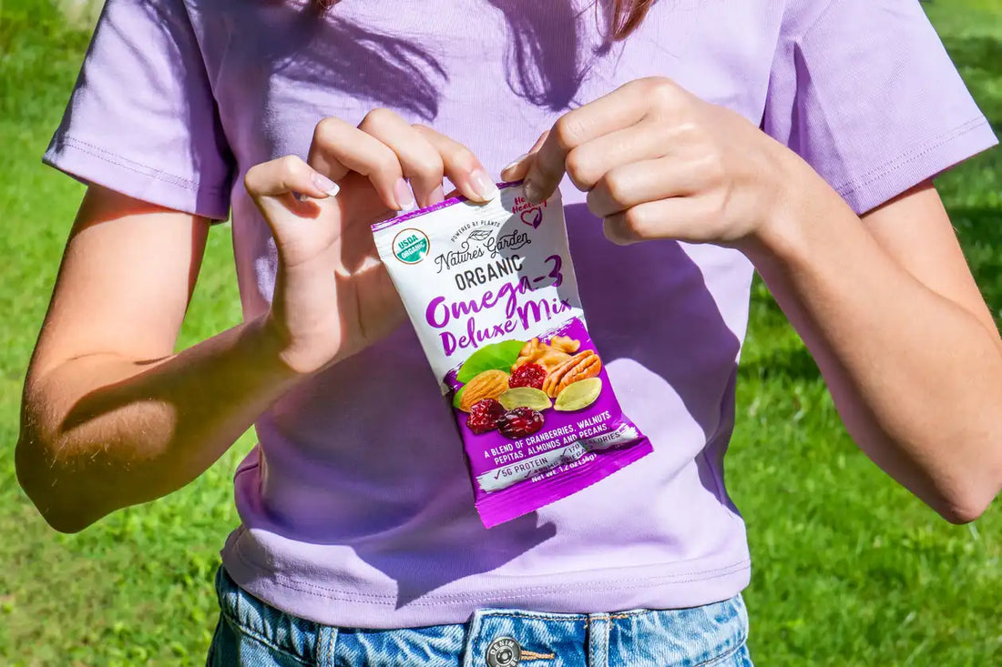 Person opening a Nature’s Garden Organic Omega-3 Deluxe Mix snack pack outdoors, featuring a healthy blend of cranberries, walnuts, almonds, pepitas, and pecans.