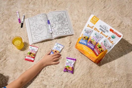 Kid grabbing Nature's Garden Organic Trail Mix Snack Pack next to coloring book, markers, and juice on plush carpet.