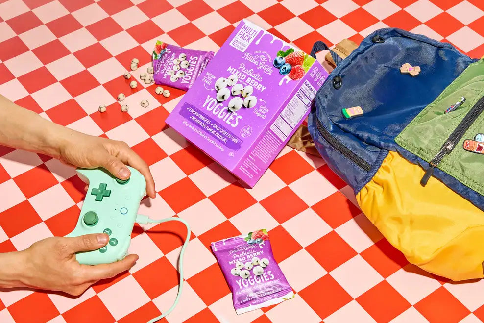 Nature’s Garden Yoggies Probiotic Mixed Berry yogurt-covered fruit snacks near backpack and game controller on red checkered table.