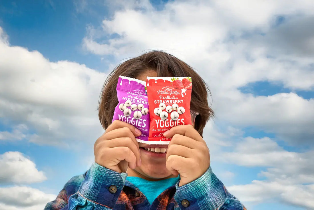 Child playfully holding Nature’s Garden Probiotic Yoggies Mixed Berry and Strawberry snack packs in front of their face against a bright, cloudy sky.