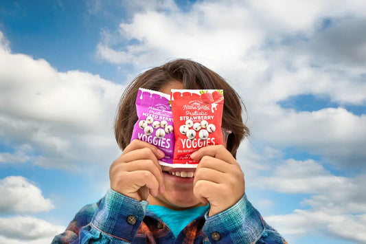 Child playfully holding Nature’s Garden Probiotic Yoggies Mixed Berry and Strawberry snack packs in front of their face against a bright, cloudy sky.
