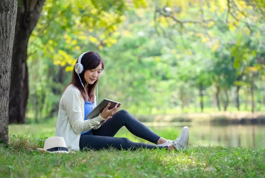 Woman relaxing with headphones and tablet by a lake, enjoying music and summer vibes—perfect pairing for Nature’s Garden snacks