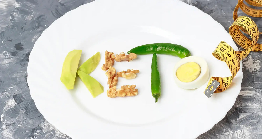 Keto spelled with avocado, walnuts, chili, and egg on a white plate beside a yellow tape measure—low-carb diet concept image.