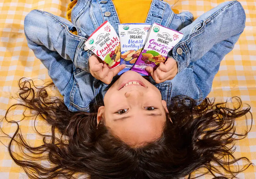 Smiling child holding Nature’s Garden Organic Trail Mix snack packs on picnic blanket, fun family snacking USA
