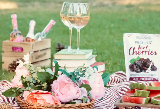 A summer picnic scene on grass featuring Nature's Garden Probiotic Tart Cherries, a wicker basket of pink flowers, fresh watermelon slices on a striped blanket, a stack of books with two glasses of white wine, and a wooden crate holding bottled drinks.