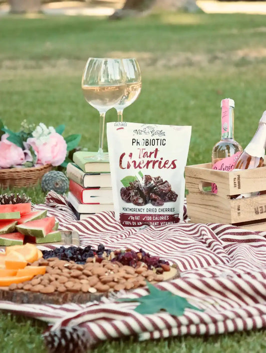 Nature’s Garden Probiotic Tart Cherries featured in an outdoor picnic setup with fruits, wine, books, and a striped picnic blanket