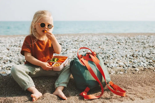 Young child eating healthy vegan snacks from a lunchbox while sitting on a pebbled beach with a backpack nearby.
