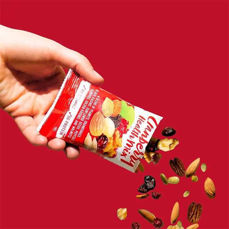 Hand holding a Cranberry Delight trail mix packet with cranberries, raisins, almonds, cashews, and pumpkin seeds spilling out on a red background.