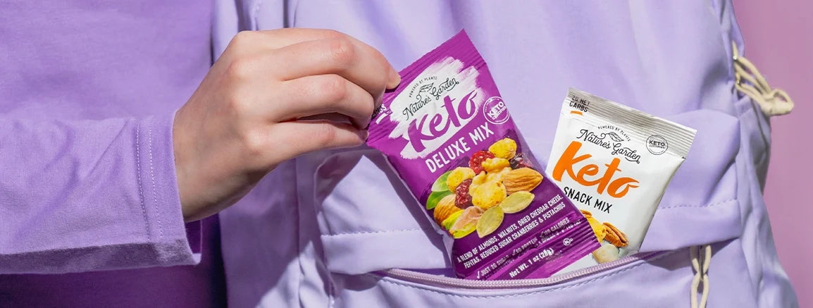 Hand placing Nature’s Garden Keto Deluxe Mix into a lavender backpack pocket alongside another low-carb, low-sugar keto snack pack.