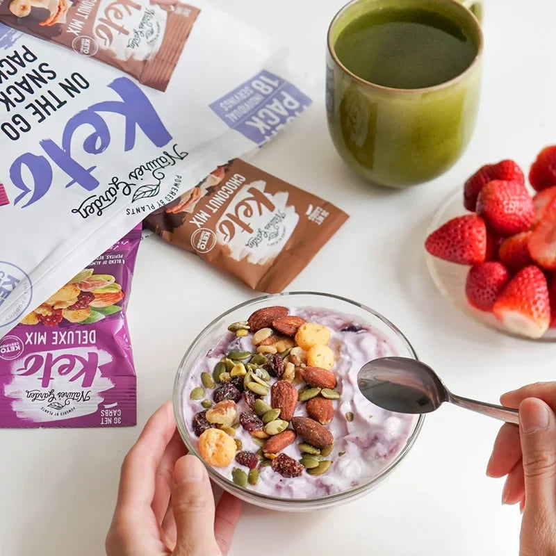 Nature’s Garden Keto Snack Mix used as a yogurt topping with strawberries and tea, featuring Choconut and Deluxe Mix packs in the background.